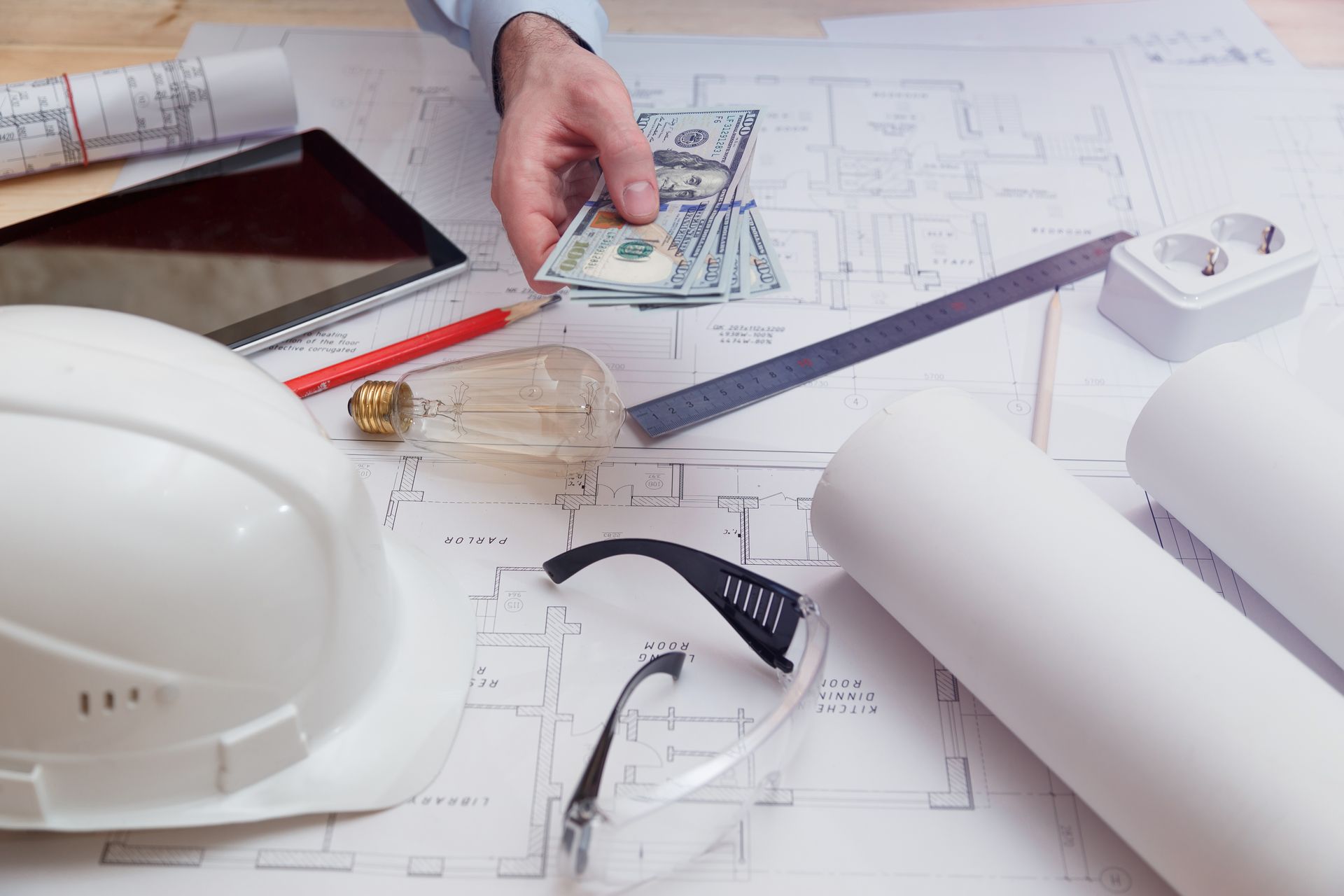 Construction plans with hard hat, tape measure, pen, blueprint tube, and a hand holding dollar bills