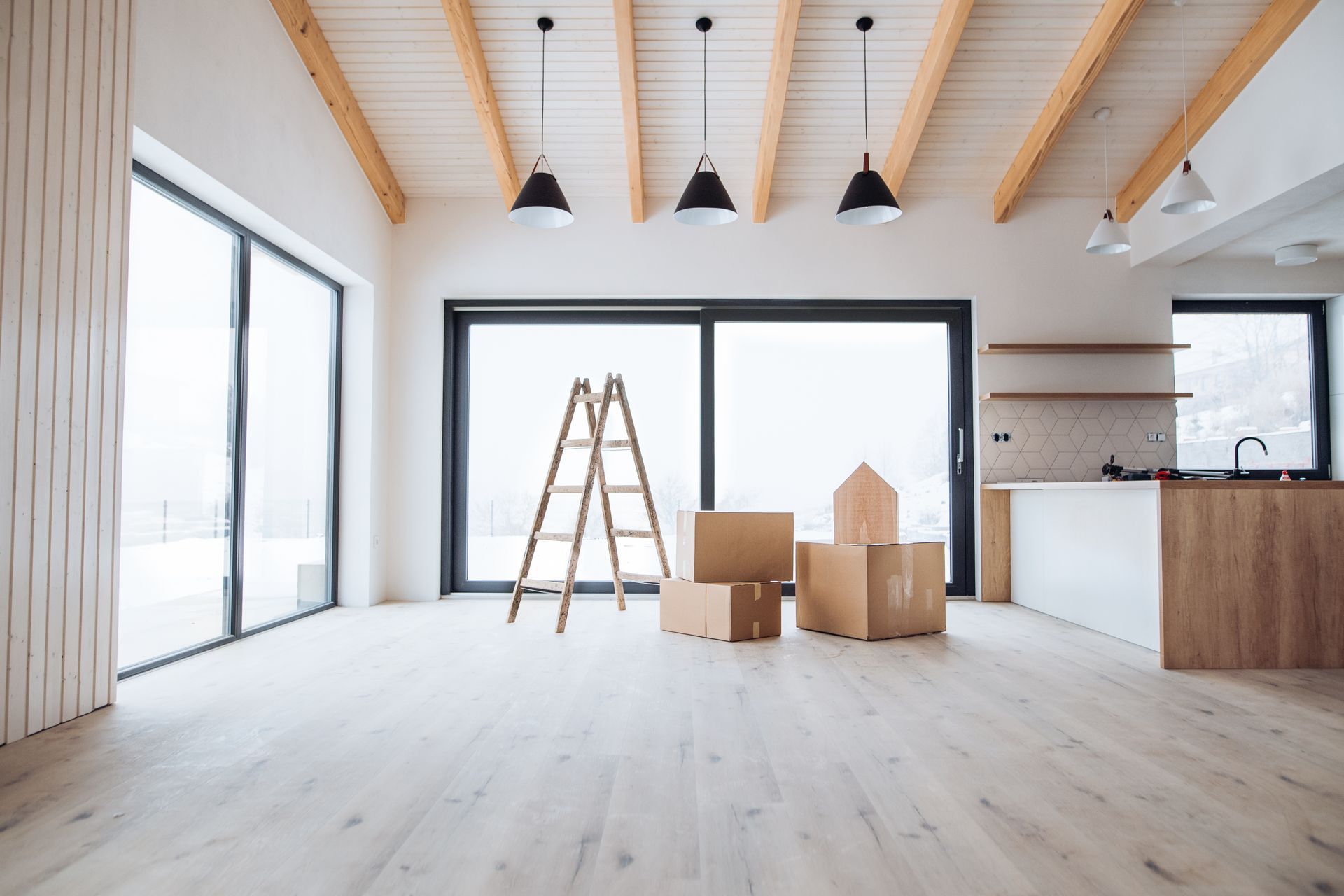 Bright empty room with ladder, moving boxes, large windows, and exposed wood ceiling