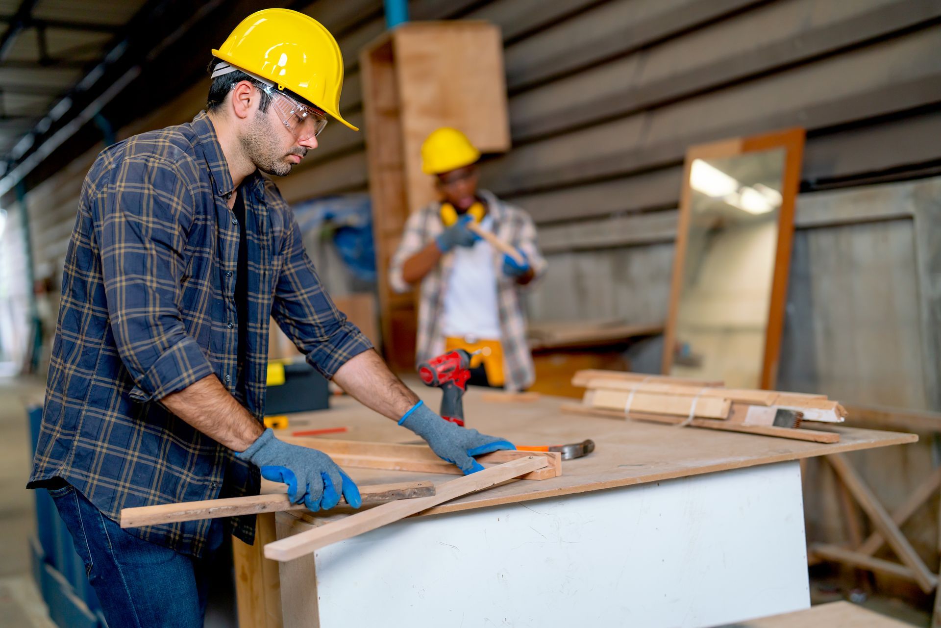 Worker in a yellow hard hat measuring a wooden board in a workshop, with another worker in the background
