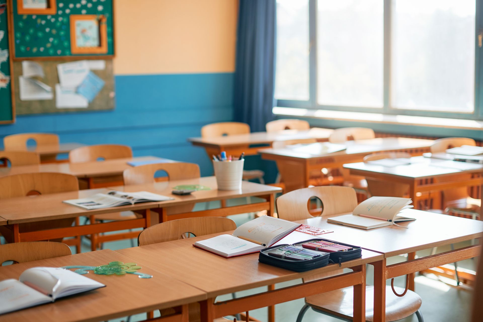 Empty classroom with wooden desks, open notebooks, and sunlight through large windows