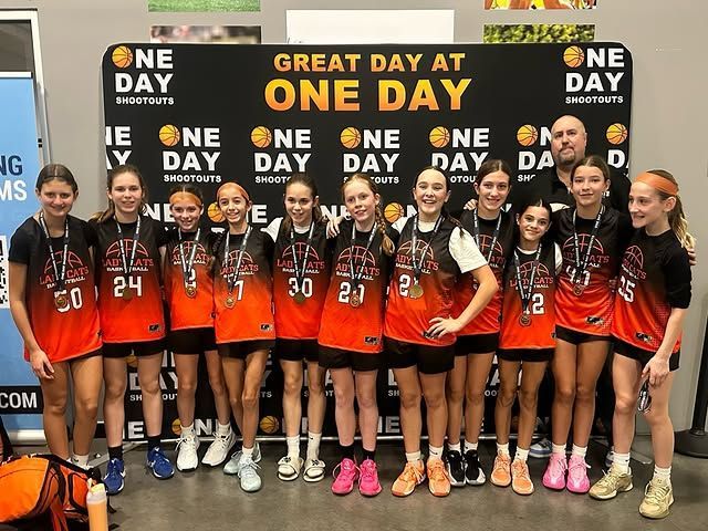 Girls basketball team posing in orange uniforms at a “Great Day at One Day” event backdrop