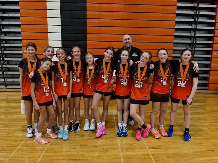 Youth basketball team posing in orange jerseys on a gym court, holding medals with a coach behind them