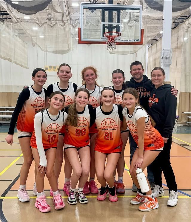 Youth basketball team posing on an indoor court in orange-and-white uniforms under the hoop