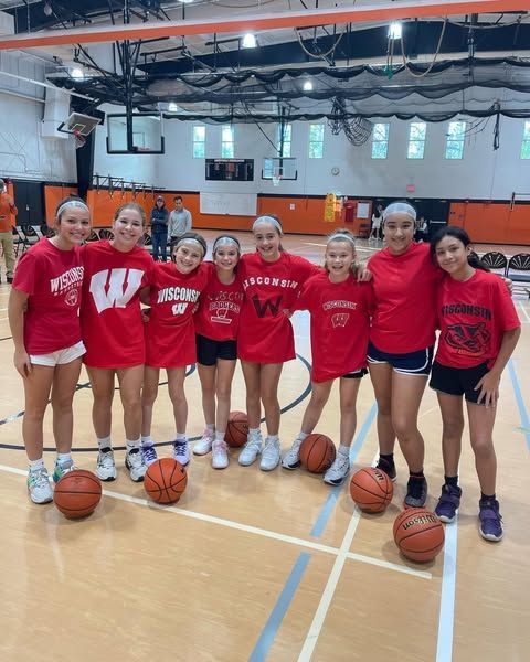 Seven people in red shirts posing with basketballs on an indoor court under gym lights