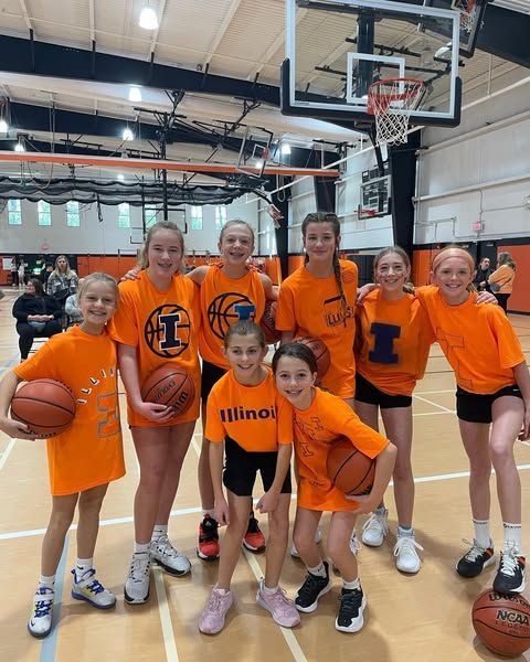 Girls’ basketball team in orange shirts posing in a gym with basketballs and a hoop
