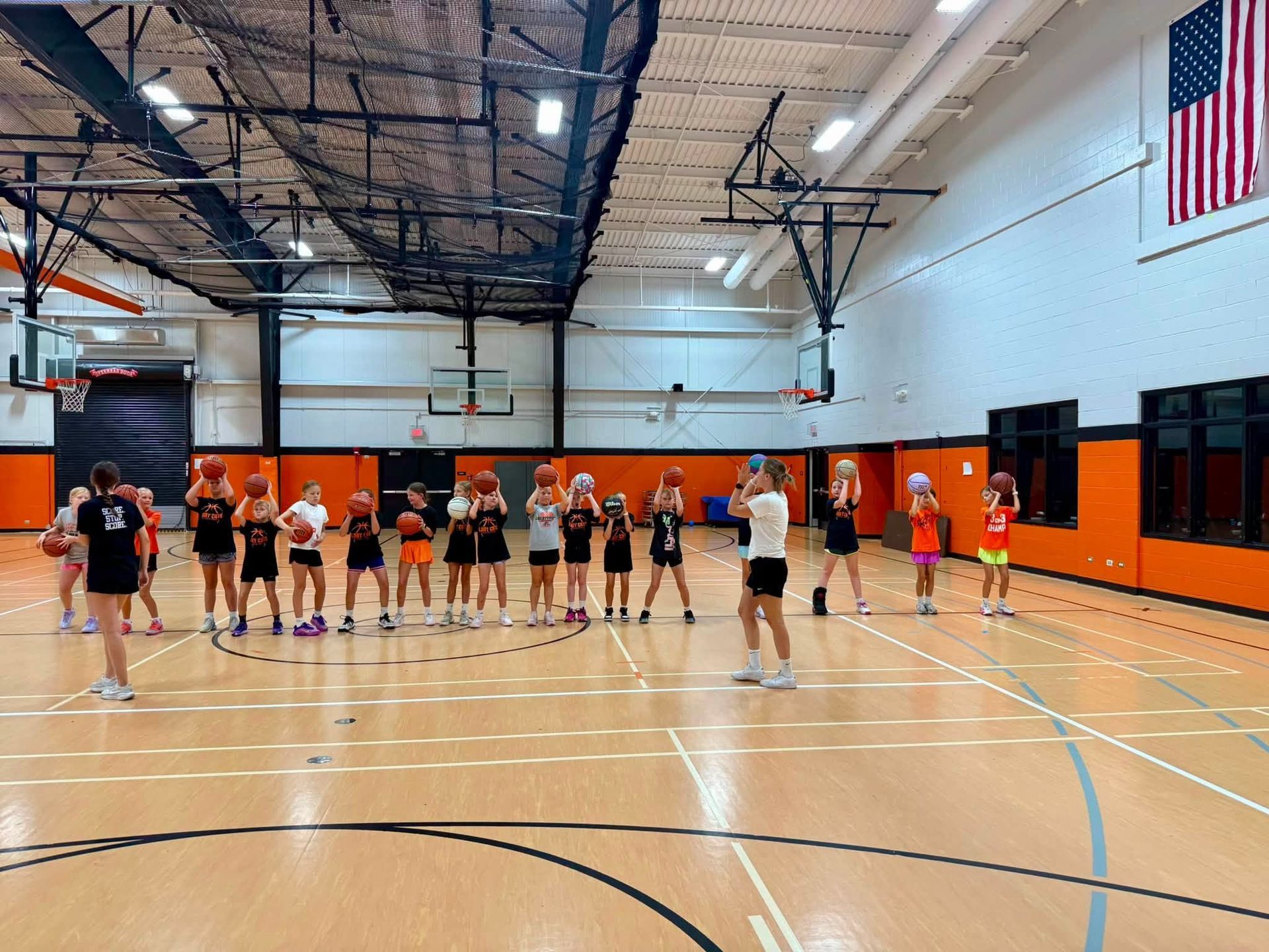 basketball team lined up on an indoor gym court under orange walls and an American flag