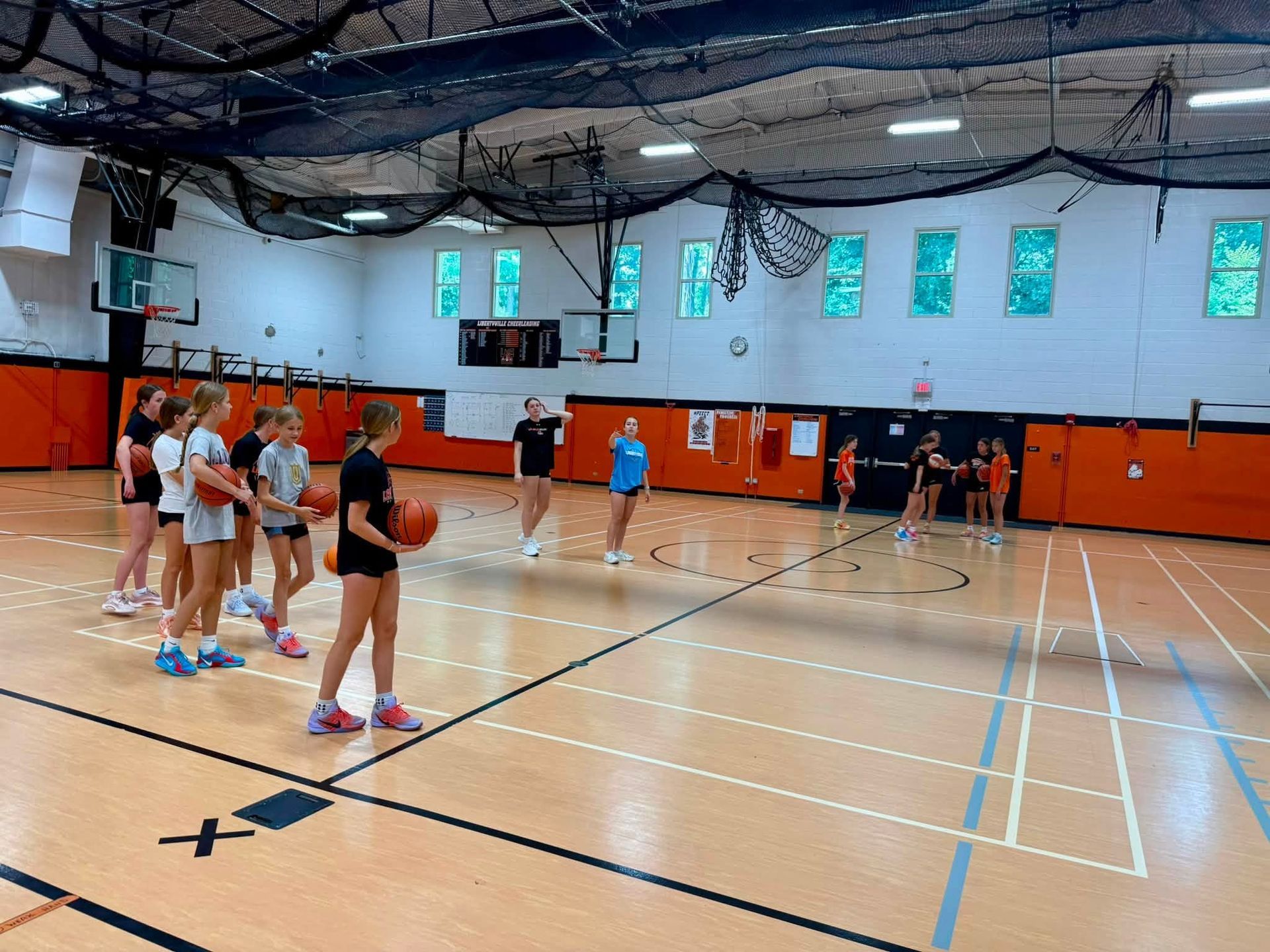 Children practicing basketball in a gym, with players lined up on a hardwood court and orange wall padding.