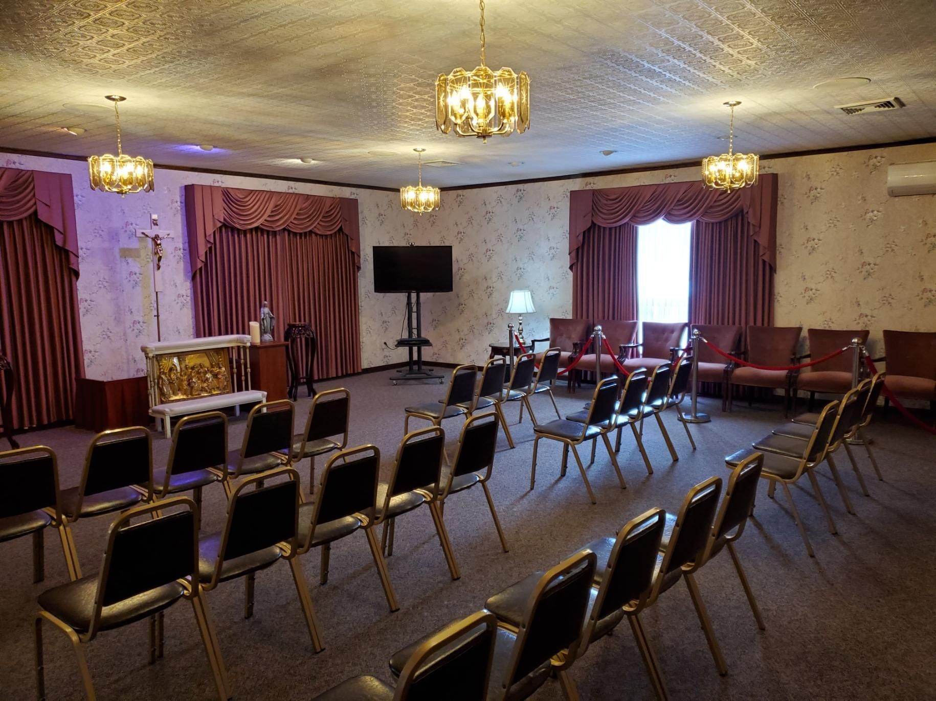 Empty meeting room with rows of chairs, curtains, chandeliers, and a TV screen on a stand.