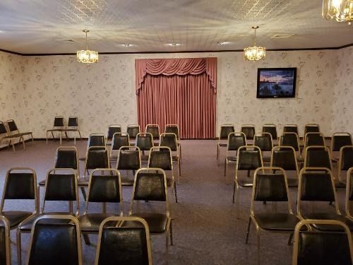 Empty banquet hall with rows of chairs, pink curtains on a stage, and a wall-mounted TV on the right.