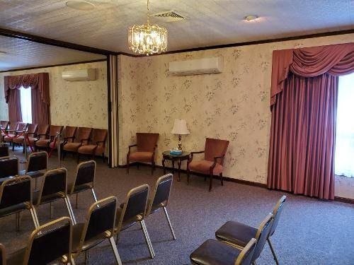 Empty meeting room with rows of chairs, floral wallpaper, chandeliers, and pink curtains over windows.