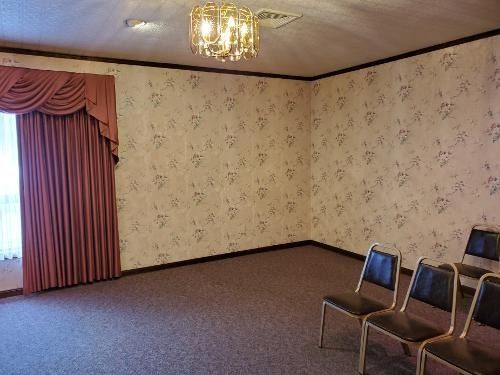 Empty carpeted room with floral wallpaper, curtained window, chandelier, and chairs along the right wall