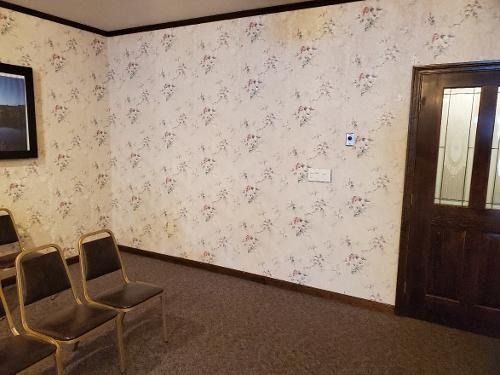 Empty waiting room with floral wallpaper, brown chairs, a wooden door, and a small wall control panel.