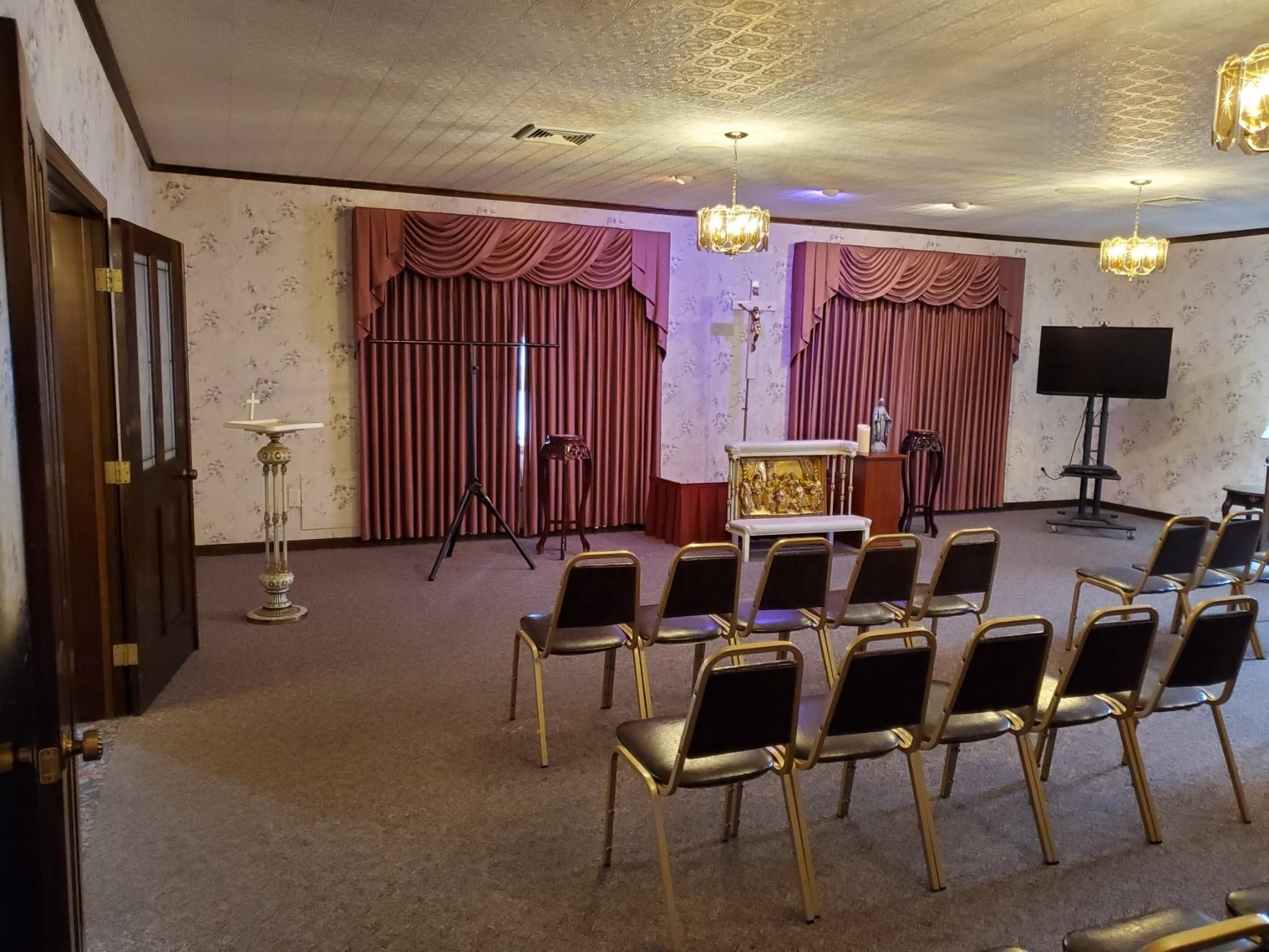 Small event room with rows of chairs facing a decorated table, red curtains, and a wall-mounted TV.