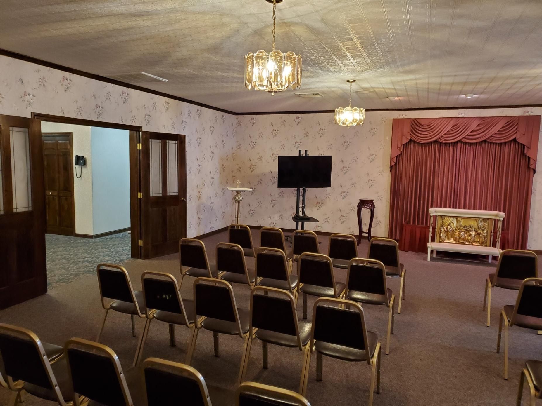 Small meeting room with rows of chairs, TV on a stand, and a stage with red curtains and a floral backdrop.