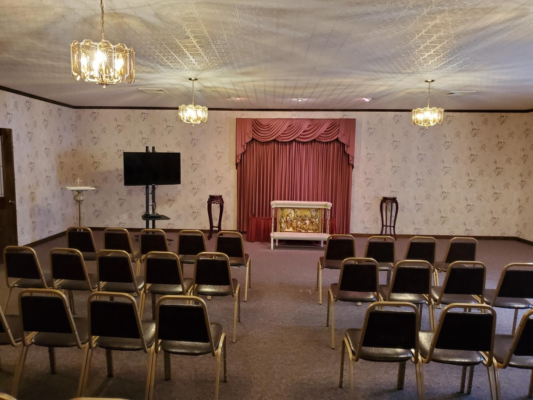 Empty church hall with rows of chairs, a small stage, red curtains, and chandeliers
