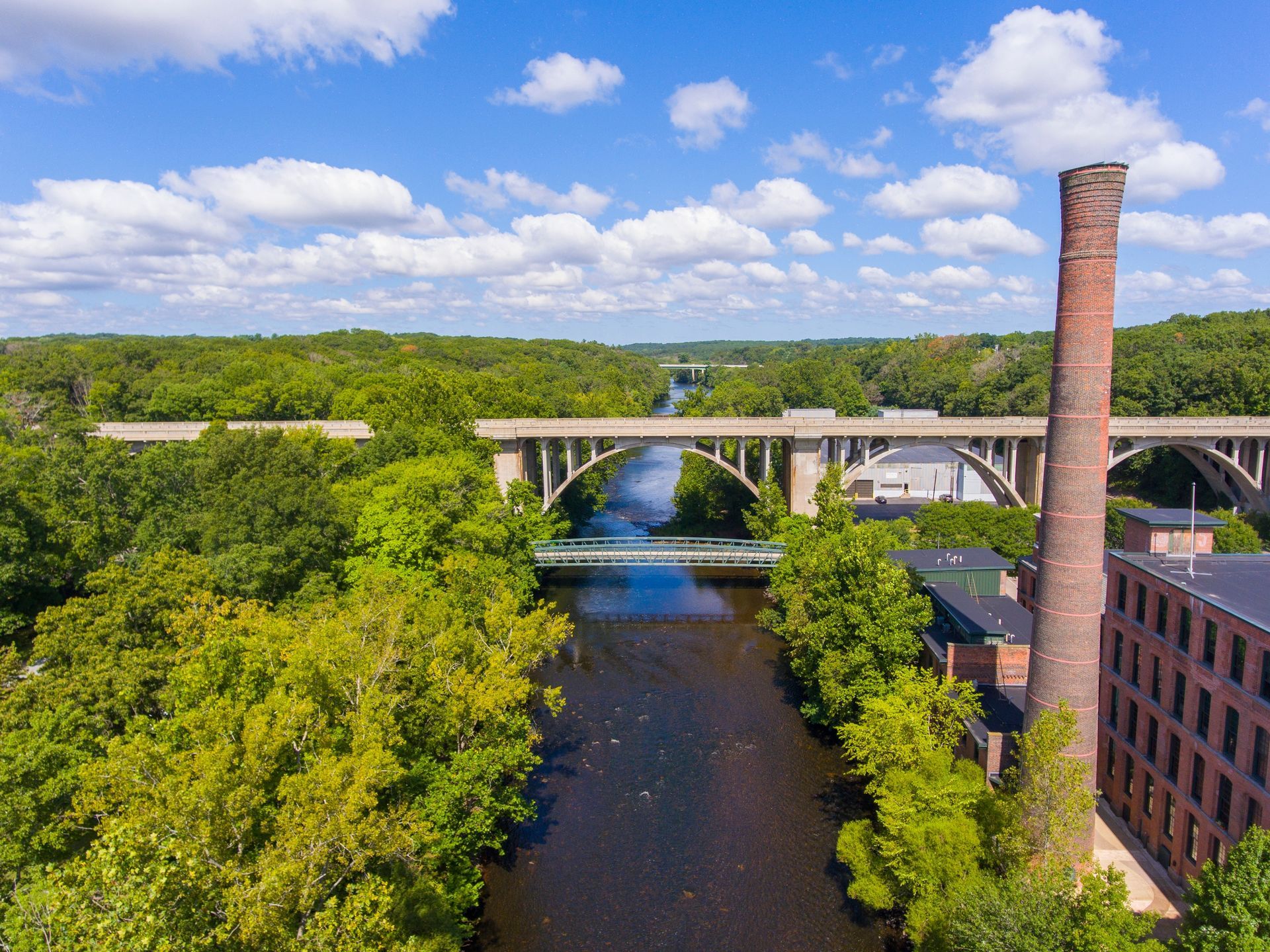 River view with a concrete bridge, brick smokestack, and lush green trees under a blue sky