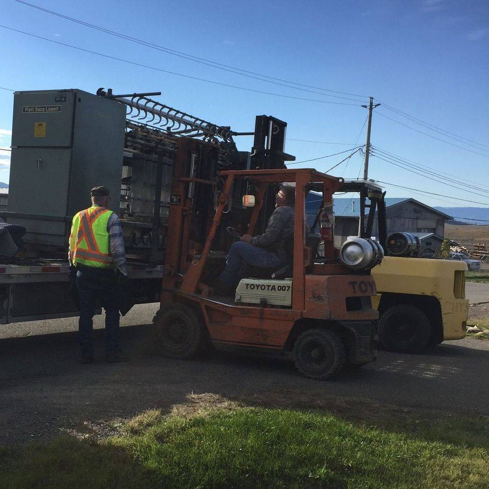 Two forklifts unloading a large metal structure from a flatbed truck; a worker in a safety vest stands nearby.