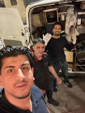 Tre persone in posa accanto a un furgone aperto di notte, una si scatta un selfie e un'altra saluta con la mano.