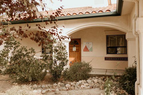 Adobe-style house entrance with archway, wooden door, and desert shrubs in front