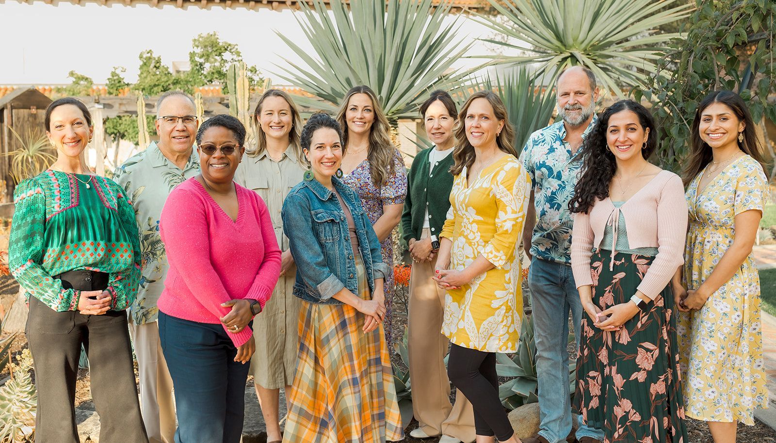 Group of nine people standing outdoors, smiling in colorful outfits with spiky desert plants behind them