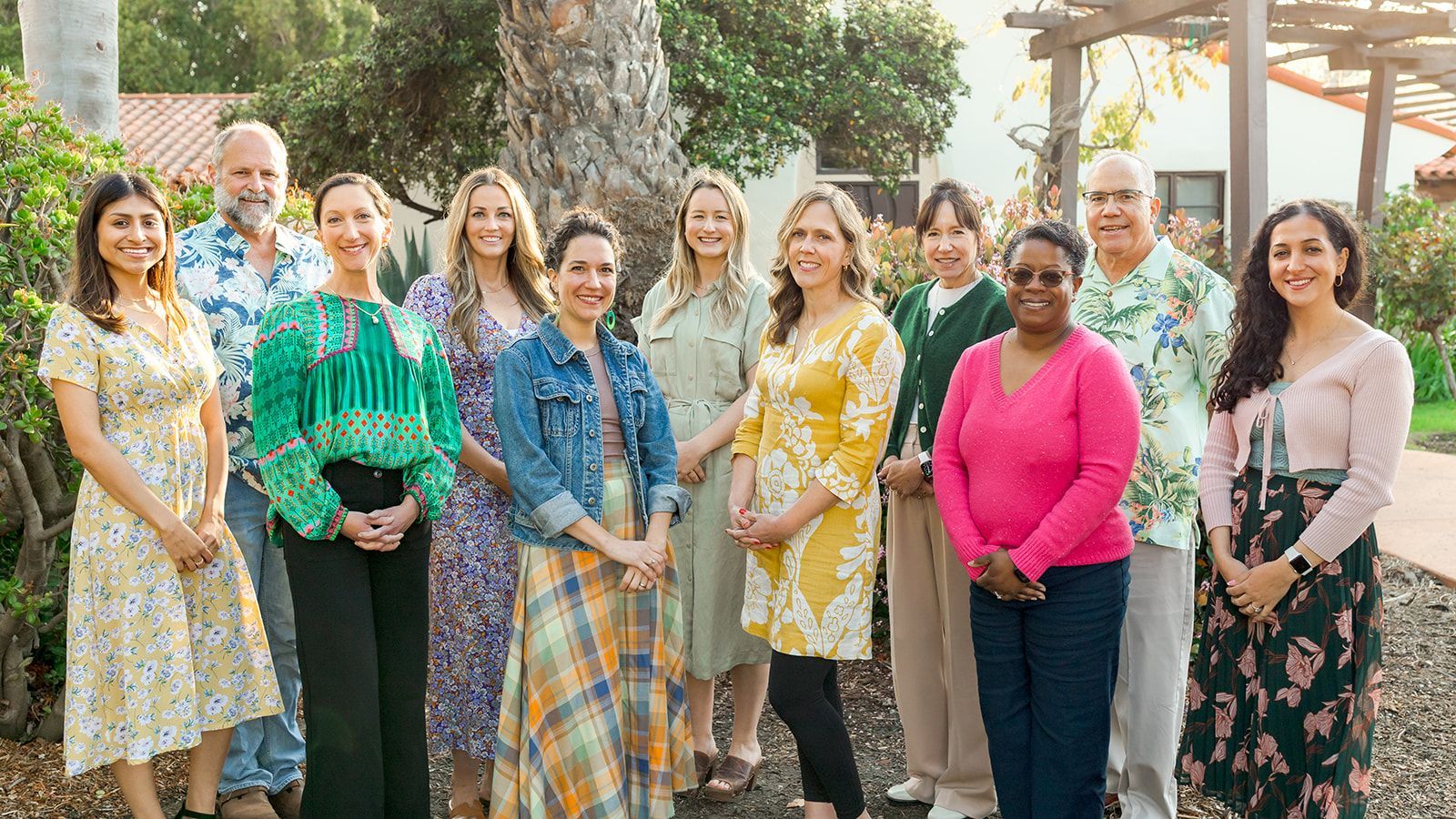 Group of people standing outdoors in a garden, smiling and dressed in colorful attire.