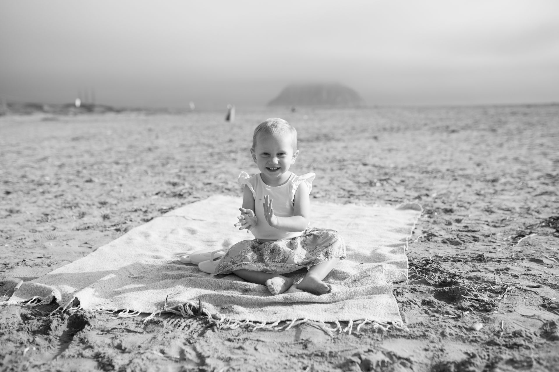 Smiling child sitting on a blanket on a sandy beach, holding a small toy.