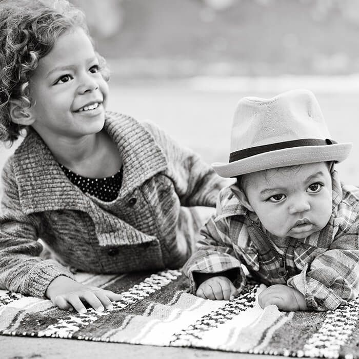 Two children lying on a blanket, smiling outdoors; one wears a hat and plaid shirt.