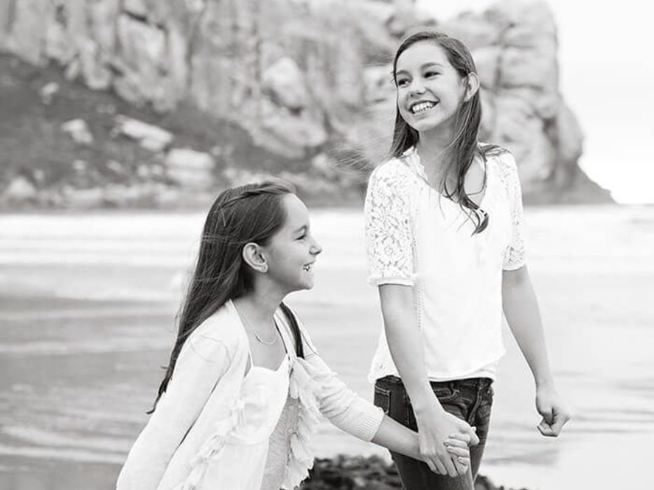 Two individuals walk hand-in-hand along a sandy beach near large rock formations.