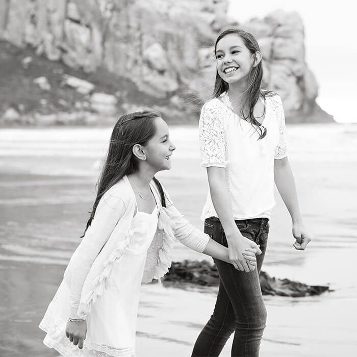 Two women holding hands and walking on a beach, smiling near the shoreline.