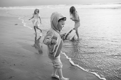 Three children play at the shoreline, one in a hooded jacket walking through wet sand.