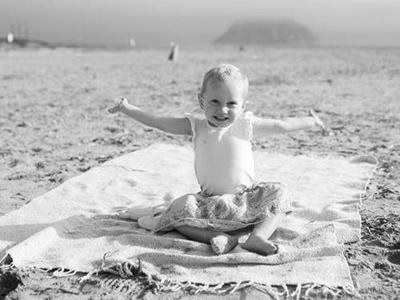 Smiling baby sitting on a blanket at the beach with arms outstretched, ocean and distant island behind.