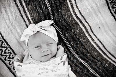 Newborn wrapped in a patterned blanket, wearing a bow headband and winking on a striped blanket.