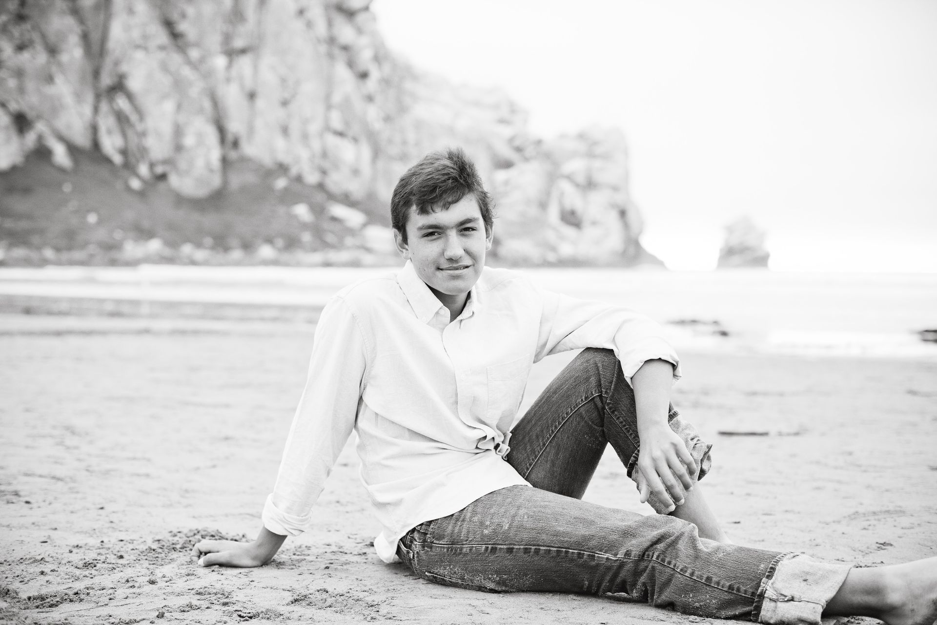 Person sitting on a beach with cliffs in the background, wearing a light shirt and dark pants.
