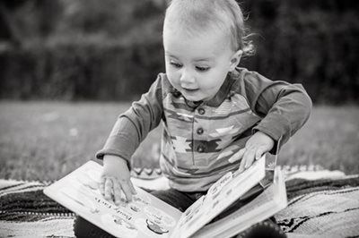 Child reading an open book on a blanket outdoors, reaching toward the pages.