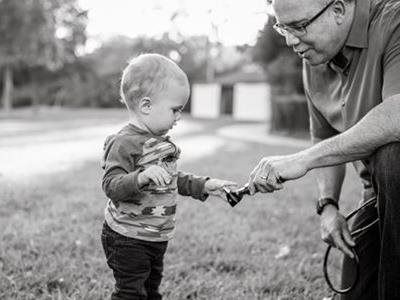 Older man offering a small plant to a toddler outdoors on a grassy lawn