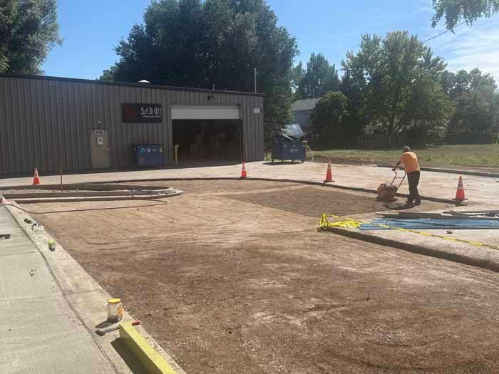 Person operating a compactor on a dirt surface outside a building, with orange cones and caution tape.