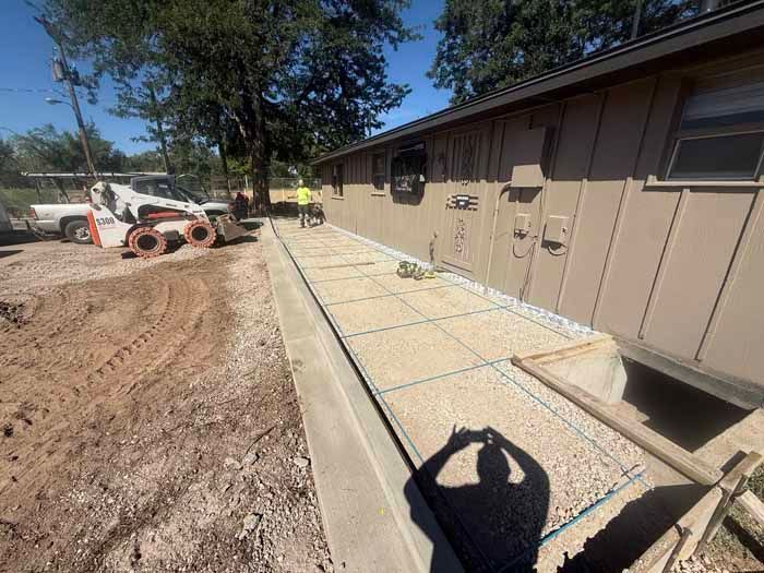 Construction site: concrete foundation being poured along the side of a building; person in a yellow vest works near a small machine.