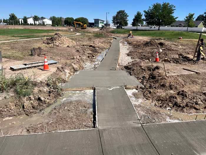 Construction of a concrete pathway in a dirt field. Workers and heavy machinery are present.