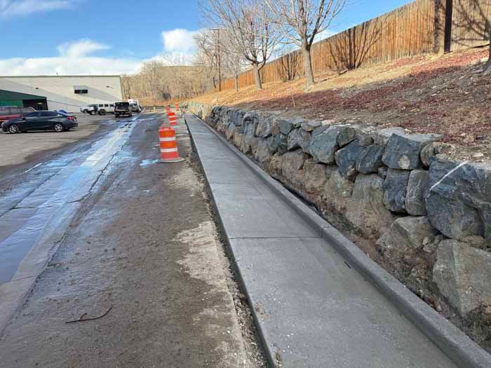 Concrete walkway next to a stone retaining wall and a sloped driveway. Cars parked in the distance.
