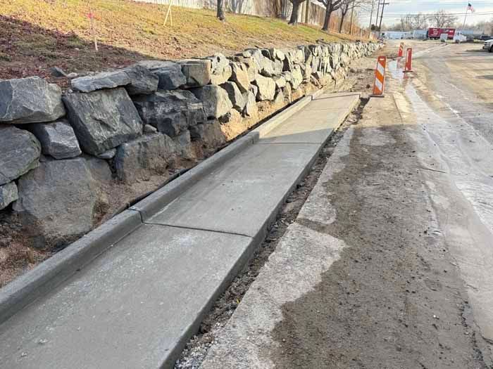 Newly poured concrete sidewalk next to a stone retaining wall and a street.