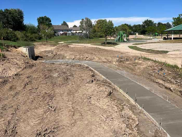 Construction site: newly poured concrete sidewalk path, dirt ground, with playground in the background.