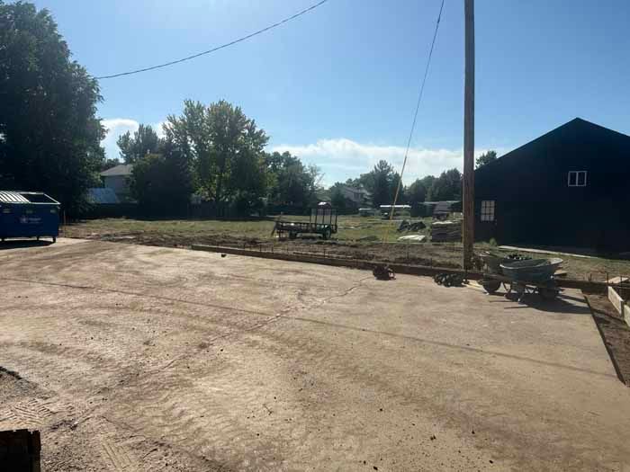 Dirt and concrete lot with a building and trailer in the background under a blue sky.