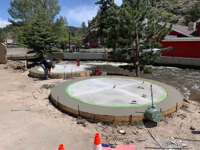 Two concrete platforms under construction near a river, with a worker, trees, and safety cones.
