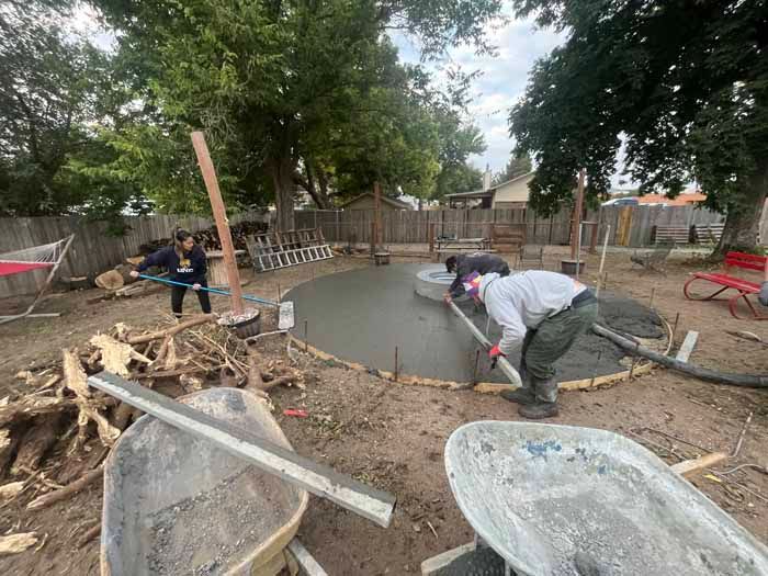 Workers pouring concrete for a circular patio in a backyard setting with trees and wheelbarrows.