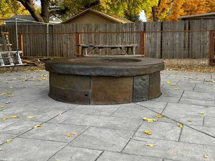 Stone fire pit on a gray patio with a wooden fence and fall foliage in the background.
