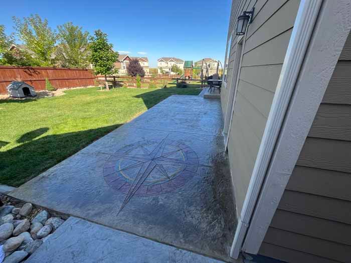 Concrete patio with compass design, beside a beige house, overlooking a grassy yard.
