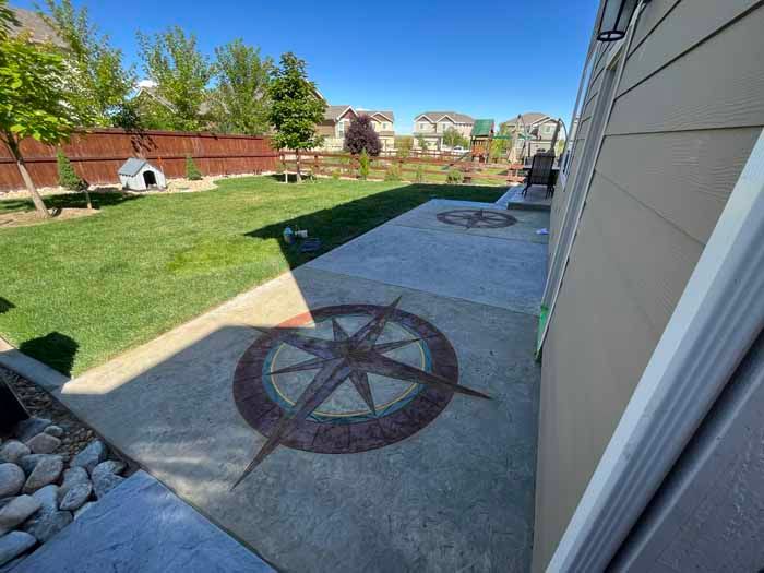 Backyard with concrete patio featuring compass rose designs, lawn, and fence.
