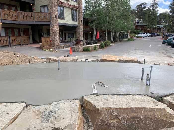 Freshly poured concrete pathway with protruding pipes, in front of a stone-faced building.