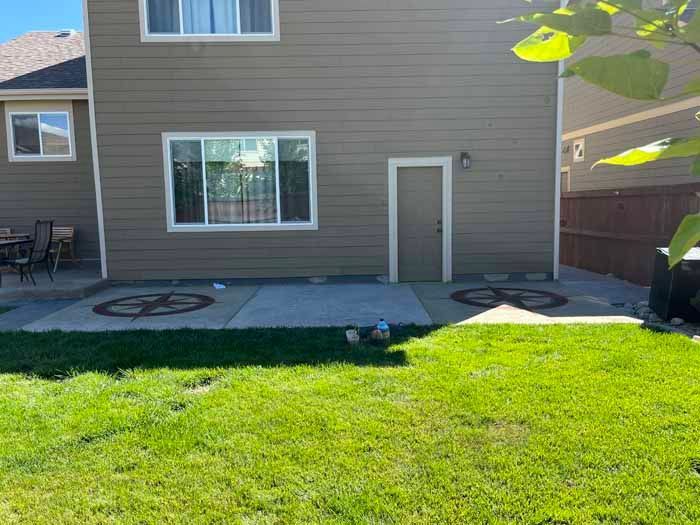 Backyard with green grass, concrete patio, tan house, door, window, and decorative fire pits.