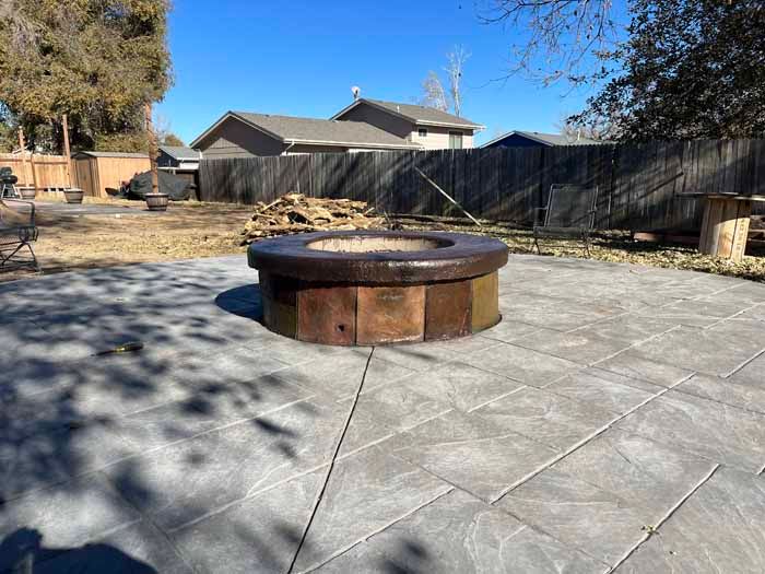 Fire pit on a gray patio in a backyard with a wooden fence and stacked firewood.